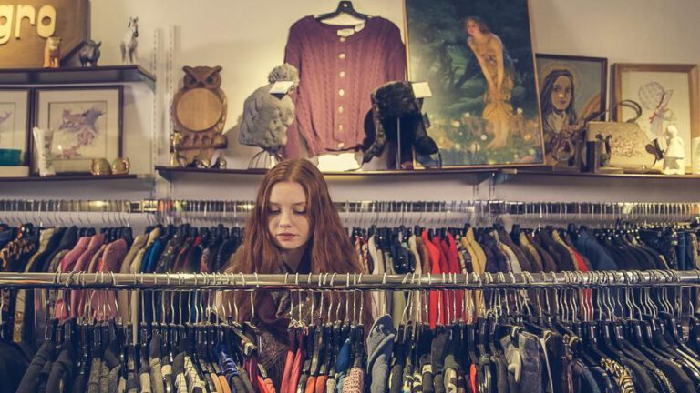 Frau in Vintage-Boutique zwischen dicht besetzten Kleiderstangen, im Hintergrund Regale mit Second-Hand-Mode und Dekorationsobjekten.