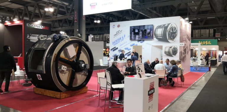 Messestand mit großem schwarzem Industrierad und Besuchern an Ausstellungsständen in Messehalle