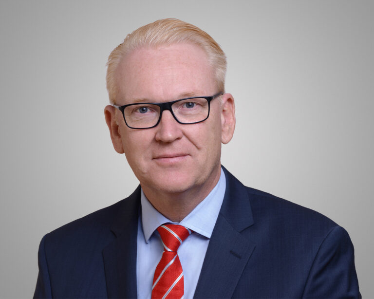 Porträt eines Mannes mit blondem Haar und Brille, dunklem Anzug und roter Krawatte vor grauem Hintergrund.