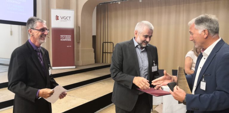 Drei Männer in Business-Kleidung stehen in einem Veranstaltungsraum. Der Mann in der Mitte hält ein burgunderrotes Objekt in den Händen, die beiden an