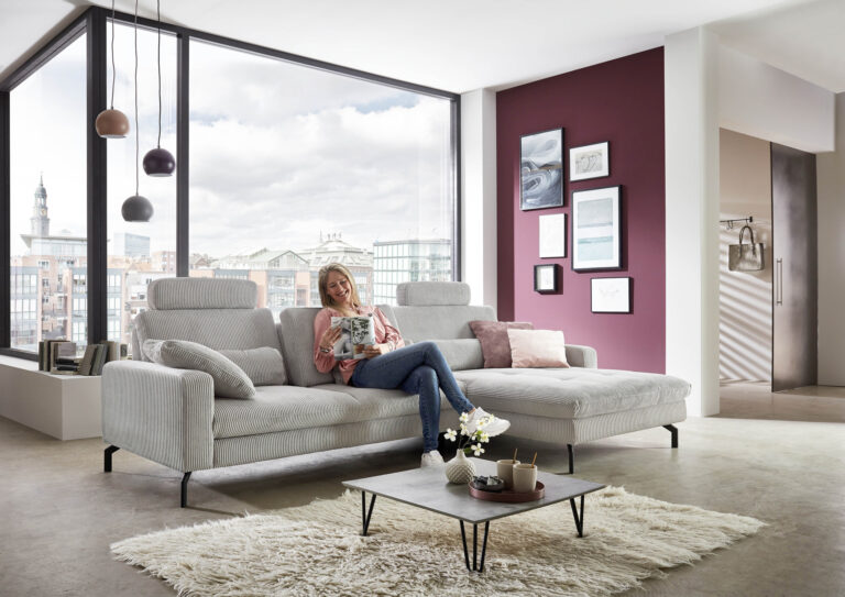 Modernes Wohnzimmer mit grauem Polstersofa, Frau sitzt auf der Couch, großes Fenster mit Stadtblick, bordeauxrote Akzentwand, Couchtisch, Pendelleucht