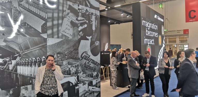 Besucher auf der Techtextil 2026 an einem Messestand mit Ausstellungswand und Schriftzug "Imagination becomes impact".