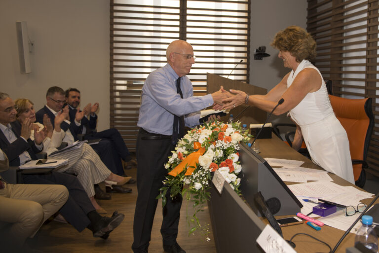Mann in blauem Hemd überreicht einer Frau in weißem Kleid eine Blumenvase, während Kollegen applaudierend zusehen.