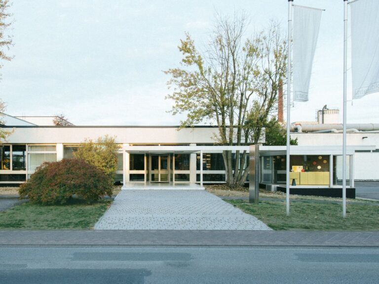 Modernes flaches Gebäude mit Glasfronten, gepflastertem Vorplatz, Flaggen und umgeben von Grünflächen und Bäumen.
