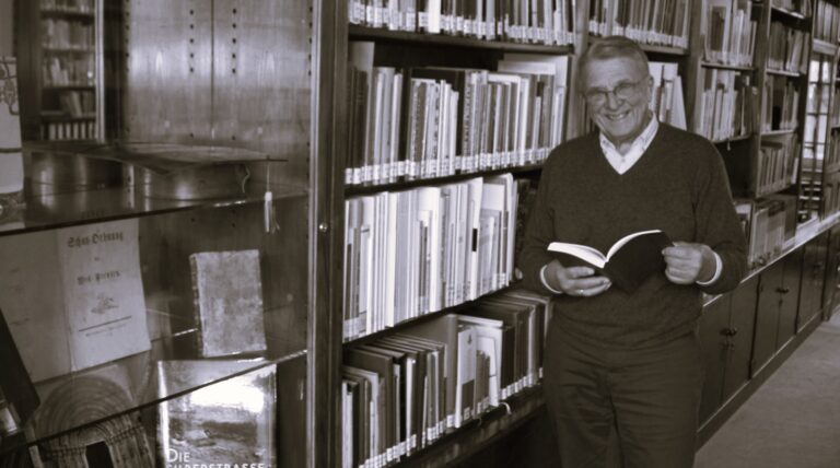 Älterer Herr mit Brille in dunklem Pullover hält offenes Buch in Bibliothek mit Bücherregalen im Hintergrund.