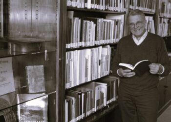 Älterer Herr mit Brille in dunklem Pullover hält offenes Buch in Bibliothek mit Bücherregalen im Hintergrund.