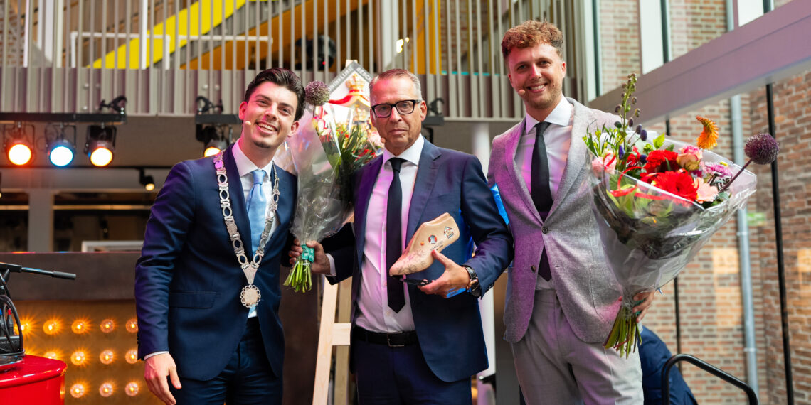 Drei Männer in Anzügen halten Blumensträuße bei einer Hoflieferanten-Ernennung, einer trägt eine Kette als Auszeichnung.